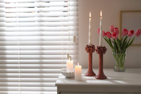 Beautiful burning candles in candlesticks and bouquet of tulips on commode at home. Space for text