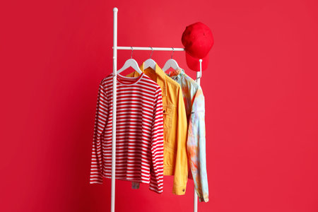 Different clothes and baseball cap hanging on rack against red background