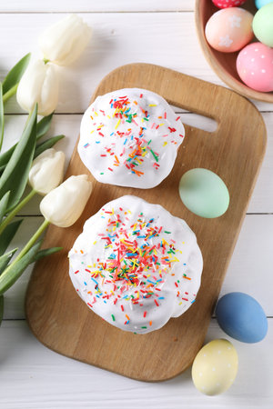 Delicious Easter cakes, painted eggs and tulips on white wooden table, flat lay