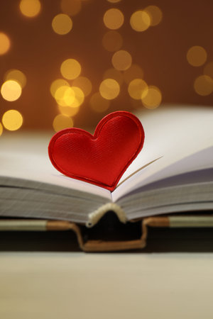 Love. Open book red decorative heart on table against blurred lights, closeup