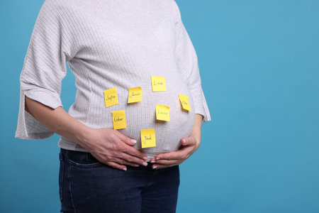 Pregnant woman with notes with different names on her belly against light blue background, closeup....