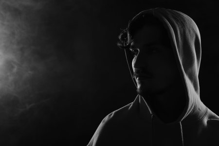 Man in hood on dark background with smoke, space for text. Black and white effect
