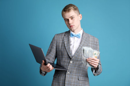 Young businessman in suit with laptop and dollar banknotes on light blue backgroundの写真素材