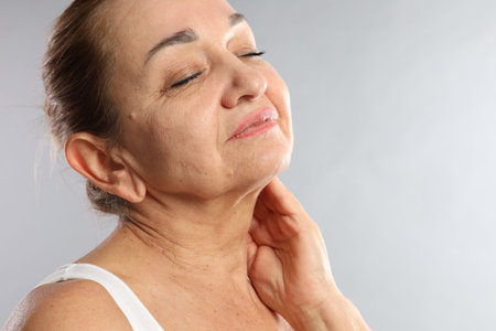 Senior woman touching her neck on light gray background, space for text