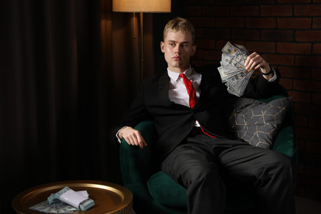 Young businessman in suit with dollar banknotes on armchair in dark room, space for text