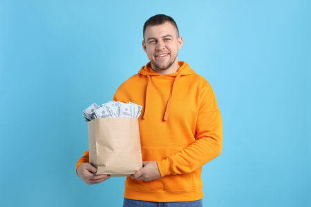 Happy man with bag full of money on light blue background
