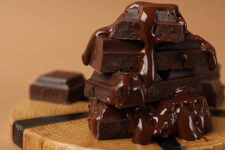 Pieces of tasty chocolate melting on wooden board against beige background, closeup