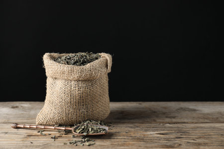 Dried green tea leaves in burlap sack and bronze scoop on wooden table against dark background. Space for textの写真素材