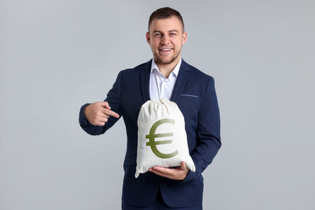 Happy businessman pointing at money bag with euro symbol on gray backgroundの写真素材