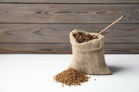 Coffee beans in sack, scoop and pile of instant coffee on white table near wooden wall, closeup. Space for textの写真素材
