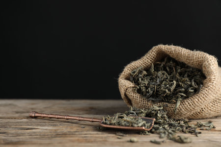 Dried green tea leaves in burlap sack and bronze scoop on wooden table against dark background, closeup. Space for textの写真素材