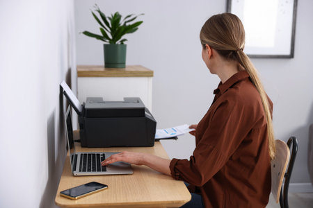 Woman taking printed sheet of paper from printer at wooden desk indoors