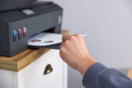 Man taking printed sheet of paper from printer at chest of drawers near grey wall indoors, closeup