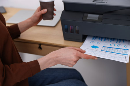 Woman taking printed sheet of paper from printer at wooden desk indoors, closeupの写真素材