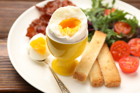 Tasty breakfast with hard boiled egg, bacon, toasted bread and salad on wooden table, closeup