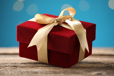 Beautiful velvet gift box on wooden table against light blue background, closeup. Bokeh effect