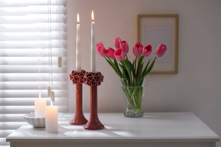 Beautiful candles in candlesticks and bouquet of tulips on commode at home