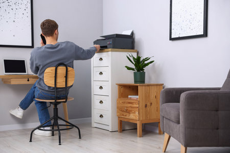 Man taking printed sheet of paper from printer and talking by smartphone indoors, back view