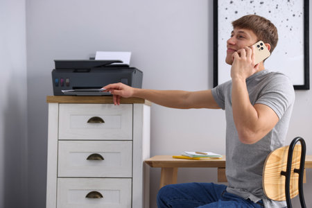 Man taking printed sheet of paper from printer and talking by smartphone indoors