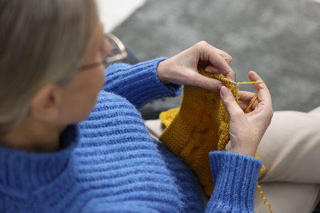 Senior woman knitting with needles on sofa at home, above view