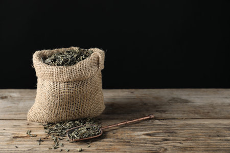 Dried green tea leaves in burlap sack and bronze scoop on wooden table against dark background. Space for textの写真素材