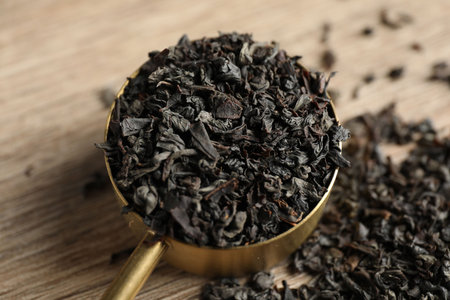 Dried black tea leaves in golden scoop on table, closeup