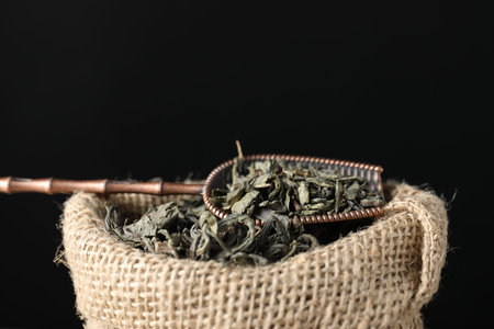 Dried green tea leaves in burlap sack and bronze scoop against dark background, closeup. Space for textの写真素材