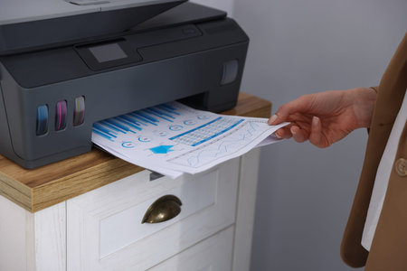 Woman taking printed sheet of paper from printer at chest of drawers indoors, closeupの写真素材