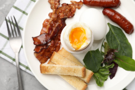 Tasty breakfast with soft boiled eggs, bacon, sausages, toasted bread and salad on gray table, top...