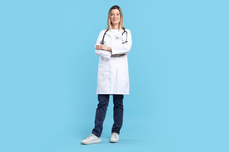 Smiling doctor in medical coat with stethoscope, badge and crossed arms on light blue background