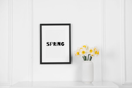 Poster with the word Spring on wall and daffodils in vase indoors