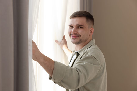 Portrait of man opening beautiful window curtains near beige wall at homeの写真素材