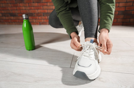 Woman tying shoelace of white sneaker indoors, closeup