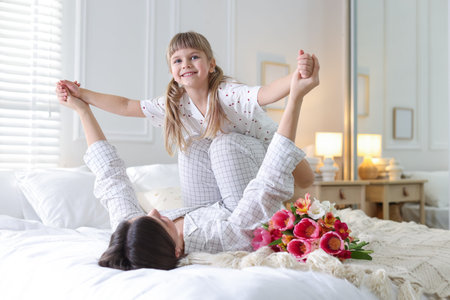 Happy Mother's Day. Cute little girl with her mother and beautiful flowers on bed at homeの写真素材