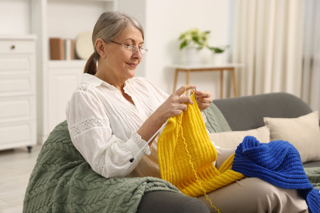 Senior woman knitting with needles on sofa at homeの写真素材