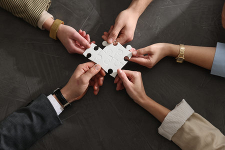 Team building. Group of businesspeople holding puzzle pieces together at dark textured table, top...