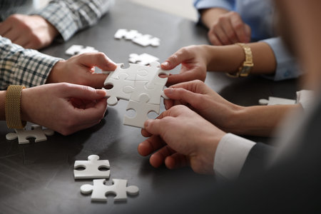 Teambuilding. Group of businesspeople holding puzzle pieces together at dark wooden table, closeup