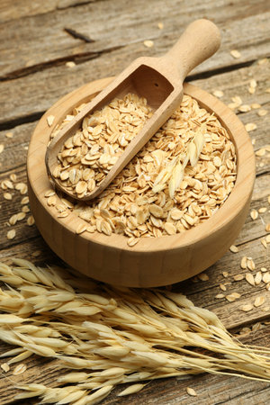 Oat flakes in bowl and panicles on wooden table, closeup