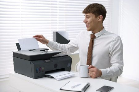 Smiling man with mug using printer at white desk in office