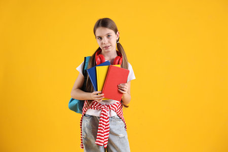 School holidays. Cute schoolgirl with backpack, books and headphones on yellow background