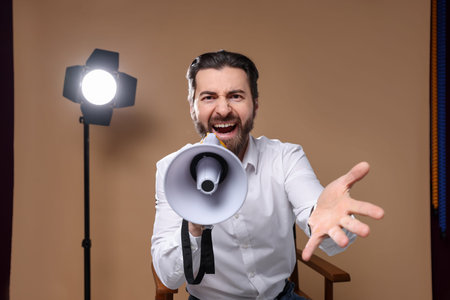 Professional film director shouting into megaphone on chair against beige background