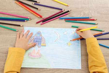 Little boy drawing with pencil at wooden table, top view