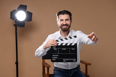 Professional film director with clapperboard pointing at something on chair against beige background