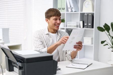 Smiling man working with document at white desk with printer in office