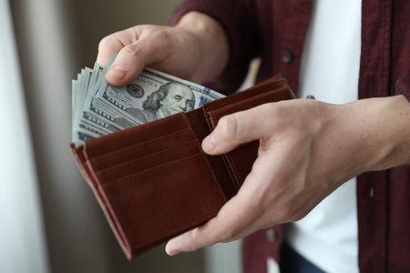 Man putting dollar banknotes into wallet against blurred background, closeup