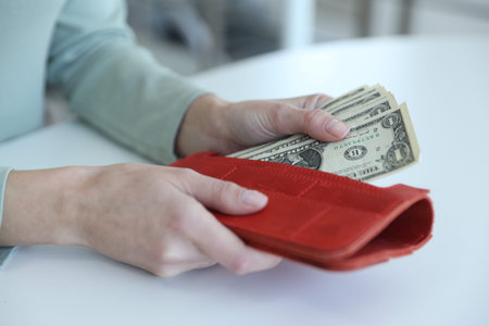 Woman putting dollar banknotes into wallet at white table, closeup