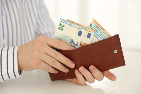 Man putting euro banknotes into wallet at white table, closeup
