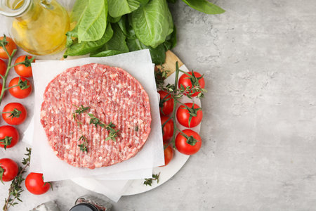Uncooked meat patty with thyme, tomatoes, lettuce and oil on gray textured table, flat lay. Space...