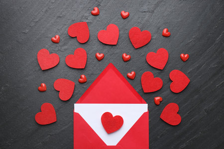 Envelope with greeting card and decorative hearts on black table, flat lay