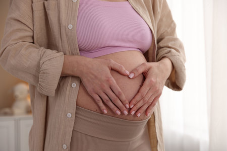 Pregnant woman making heart with hands on her belly at home, closeup
